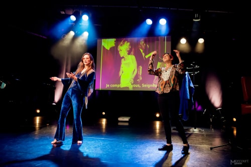 Deux personnes se tiennent sur une scène éclairée, avec un écran en arrière-plan montrant une image et du texte en français, dans un environnement de spectacle ou de théâtre.