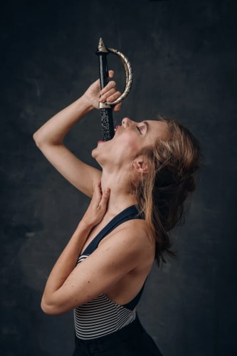 Femme tenant une épée près de sa bouche, simulant un acte dramatique, fond sombre.