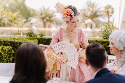 Une femme vêtue d'une robe d'époque rose et jaune avec un éventail dentelé blanche, portant un collier en dentelle et une coiffe fleurie, souriant à deux personnes assises à une table, dans un jardin avec des palmiers en arrière-plan.