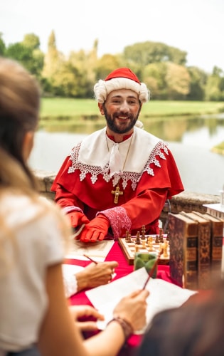 Homme vêtu d'un costume historique rouge et blanc, avec une croix dorée autour du cou, assis à une table en plein air près de l'eau, souriant et discutant avec deux personnes tenant des papiers et des crayons, avec un jeu d'échecs et des livres anciens sur la table.