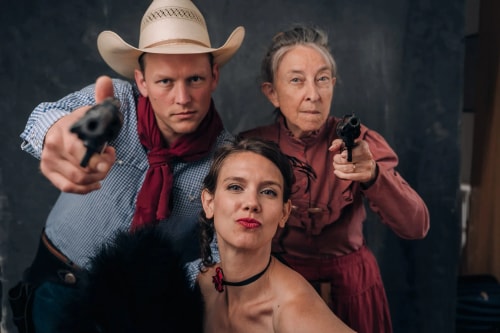 Trois personnes déguisées en style western avec un homme portant un chapeau de cowboy et un foulard rouge tenant un revolver pointé vers la caméra, une femme âgée en robe rouge tenant aussi un revolver, et une jeune femme au premier plan avec un collier noir et un maquillage marqué.