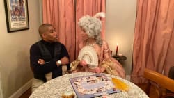 Deux personnes assises à une table ronde avec une nappe en dentelle, jouant à un jeu de société ; une personne porte un costume d'époque avec une perruque blanche et tient un éventail, l'autre personne est habillée de façon contemporaine avec un pull noir et une montre.