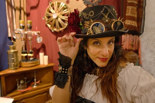 Frances Vieras-Blanc portant un chapeau steampunk orné de rouages et de lunettes, vêtue d'une blouse blanche et d'un bracelet noir, dans un décor vintage avec des objets anciens en arrière-plan.