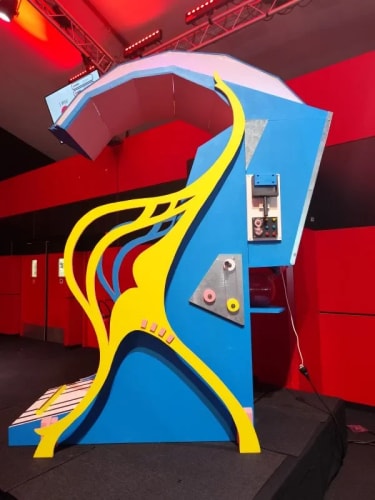 structure artistique abstraite en bleu et jaune avec des formes ondulées, placée dans une pièce avec des murs rouges et un sol noir