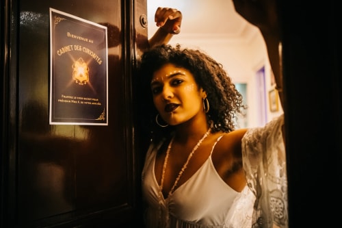 Femme aux cheveux bouclés et maquillage tribal s'appuyant contre une porte en bois foncé avec une affiche indiquant "Bienvenue au Cabinet des Curiosités".