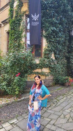 Une femme vêtue d'une robe bleue fleurie et d'un châle bleu pose devant un bâtiment ancien recouvert de lierre, avec une bannière noire portant un logo et le texte "The College of Extraordinary Experiences".
