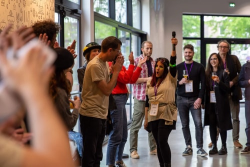 Groupe de personnes réunies dans une pièce lumineuse, certaines applaudissant, une femme au centre levant un micro en l'air, ambiance animée et conviviale.