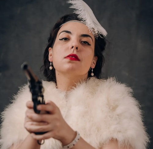 Femme avec un chapeau orné d'une plume blanche, portant un manteau en fausse fourrure blanche, tenant un pistolet pointé vers l'avant, fond sombre.