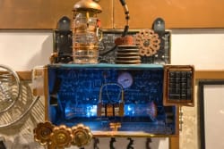 Appareil de style steampunk avec tubes à vide éclairés, engrenages, cadran de pression et clavier numérique, le tout dans un boîtier métallique avec des schémas scientifiques en arrière-plan.