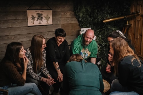 Groupe de personnes assises en cercle dans une pièce en bois, souriant et discutant ensemble.