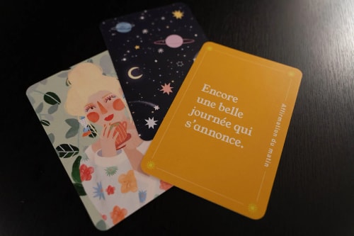 Trois cartes éparpillées sur une surface sombre, dont une carte jaune avec le texte « Encore une belle journée qui s'annonce. » et une illustration colorée d'une femme avec une tasse, ainsi qu'une carte avec un motif céleste étoilé.