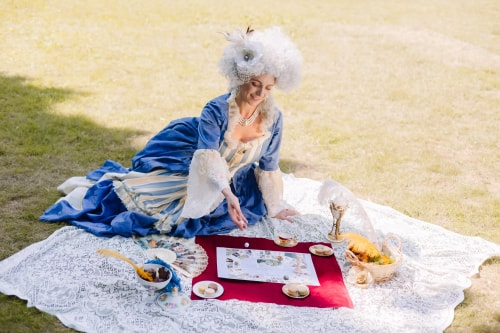 Femme en robe d'époque bleue avec perruque blanche assise sur une nappe en dentelle blanche dans un parc, jouant à un jeu de société avec des macarons, une tasse de thé, un éventail et un panier avec une plume jaune.