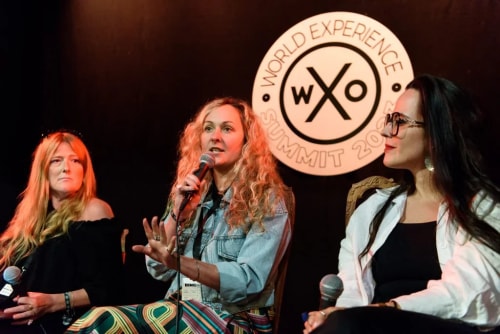 Trois femmes assises sur une scène lors d'une conférence, l'une d'elles parle dans un micro, en arrière-plan un logo avec les mots World Experience Summit 2023.