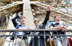 Trois hommes debout sur un balcon en métal, regardant vers le haut, avec un homme pointant du doigt, sous une structure métallique beige.