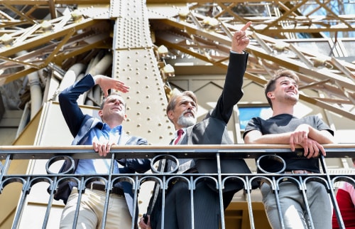 Trois hommes debout sur un balcon en métal, regardant vers le haut, avec un homme pointant du doigt, sous une structure métallique beige.