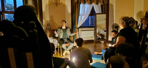 Une personne parle assise sur une chaise dans une pièce éclairée avec des spectateurs assis par terre et sur des chaises, un miroir reflète plusieurs personnes à l'arrière.