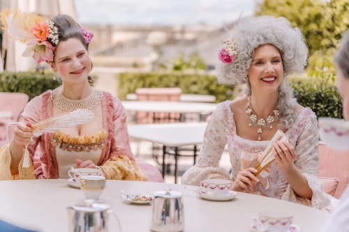 Deux femmes souriantes en costumes d'époque élégants avec des accessoires floraux tenant des éventails, assises à une table extérieure avec des tasses à thé.
