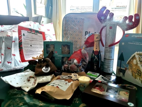 image montrant plusieurs cadeaux et objets de fête sur une table devant une fenêtre, comprenant des biscuits de Noël, une bouteille de champagne, un serre-tête avec des bois de renne, un bonbon canne, une bougie parfumée, un jeu de pâte à modeler, une maison en pain d'épice, et un sac cadeau avec motif de rennes rouges