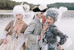 Trois personnes habillées en costumes historiques du XVIIIe siècle avec perruques blanches, debout près d'un plan d'eau, en train de discuter et de sourire.