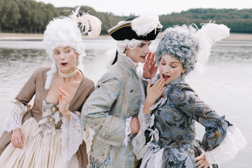 Trois personnes habillées en costumes historiques du XVIIIe siècle avec perruques blanches, debout près d'un plan d'eau, en train de discuter et de sourire.