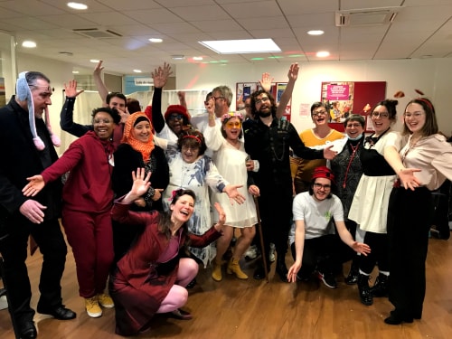 Groupe de personnes poser ensemble dans une salle, souriant et levant les mains, certains portant des accessoires festifs comme des oreilles de lapin, un foulard orange, un chapeau rouge, et des lunettes jaunes. Ambiance joyeuse et conviviale.