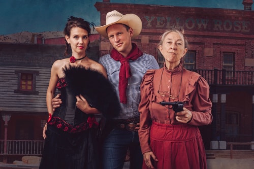 Trois personnes en costumes d'époque du Far West posent devant un bâtiment ancien; une femme porte une robe noire et rouge avec un éventail noir, un homme porte un chapeau de cowboy blanc et un foulard rouge, et une femme âgée porte une robe rouge avec un revolver dans la main.