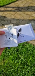 Main tenant une note avec un message en anglais et un paquet blanc marqué 'EXTRAORDINARY.CARDS' en arrière-plan, le tout sur un fond d'herbe verte et de pavés
