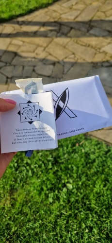Main tenant une note avec un message en anglais et un paquet blanc marqué 'EXTRAORDINARY.CARDS' en arrière-plan, le tout sur un fond d'herbe verte et de pavés