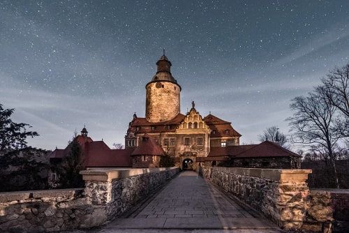 Château ancien éclairé la nuit sous un ciel étoilé avec un chemin en pierre menant à l'entrée principale.