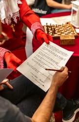 Personne en tenue rouge avec des gants rouges tenant un document écrit à la main, tandis qu'une autre personne tient un crayon rouge au-dessus du document, avec un jeu d'échecs et une lanterne blanche en arrière-plan.
