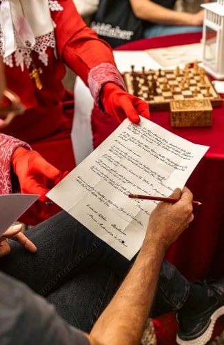 Personne en tenue rouge avec des gants rouges tenant un document écrit à la main, tandis qu'une autre personne tient un crayon rouge au-dessus du document, avec un jeu d'échecs et une lanterne blanche en arrière-plan.