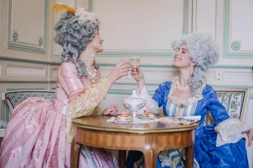 Deux femmes en costumes d'époque élégants avec des perruques grises élaborées trinquent avec des verres de vin blanc autour d'une table ronde en bois sur laquelle se trouvent des macarons et une sucrière.