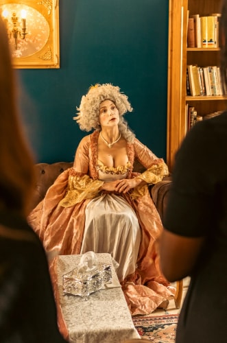 Femme habillée en costume d'époque assise sur un canapé, portant une perruque grise bouclée et un collier de perles, avec deux personnes floues au premier plan et une étagère remplie de livres en arrière-plan.