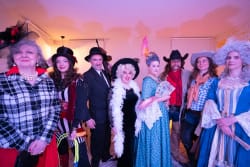 Groupe de huit personnes vêtues de costumes variés d'époques différentes, y compris des robes d'époque, des chapeaux et des accessoires comme un éventail et une plume, posant à l'intérieur devant un mur clair.