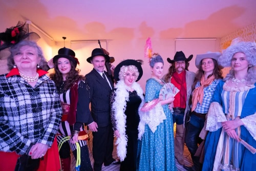 Groupe de huit personnes vêtues de costumes variés d'époques différentes, y compris des robes d'époque, des chapeaux et des accessoires comme un éventail et une plume, posant à l'intérieur devant un mur clair.