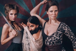 Trois femmes en costumes dramatiques posant avec confiance, l'une tient un sabre, une autre porte une étole et un bijou au front, la troisième lève le bras en signe de force.