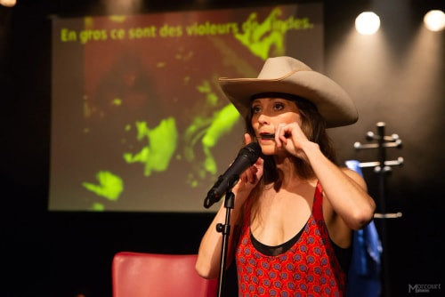 Femme portant un chapeau de cowboy et un débardeur rouge à motifs, jouant de l'harmonica devant un microphone sur scène avec un écran flou en arrière-plan.