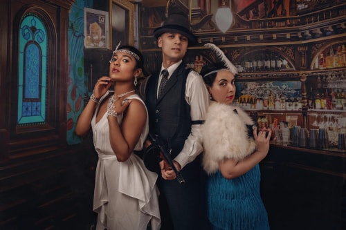 Trois personnes vêtues de costumes des années 1920 posant dans un bar vintage, une femme en robe blanche et bijoux, un homme en costume sombre avec un chapeau tenant un fusil, et une femme en robe bleue avec une étole en plumes et une plume dans les cheveux.