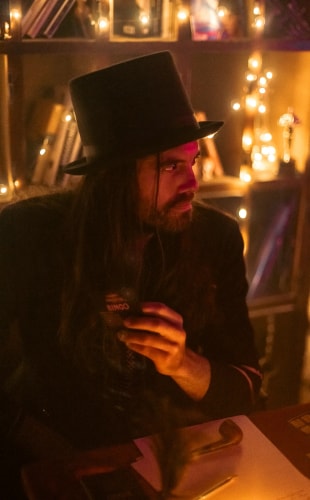 Homme aux cheveux longs et barbe portant un chapeau haut de forme noir, assis à une table dans une ambiance tamisée avec des lumières décoratives en arrière-plan, tenant une carte de bingo.