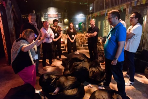 Groupe de personnes debout en cercle dans une salle avec des sacs de couchage au sol, certaines souriant et une femme gesticulant avec enthousiasme.