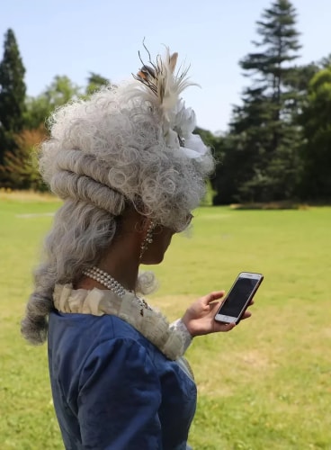 Personne vêtue d'un costume d'époque avec une perruque blanche et bouclée, observant un téléphone portable dans un parc ensoleillé.