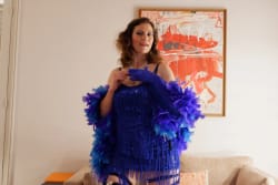 Femme vêtue d'une robe à franges bleue et d'un boa à plumes bleues et violettes, posant dans un salon avec un tableau abstrait rouge et blanc en arrière-plan.