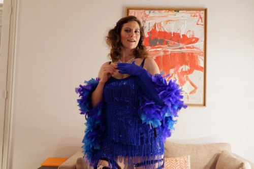 Femme vêtue d'une robe à franges bleue et d'un boa à plumes bleues et violettes, posant dans un salon avec un tableau abstrait rouge et blanc en arrière-plan.