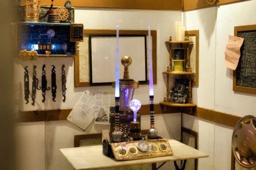 Appareil steampunk avec des tubes lumineux et des engrenages posé sur une table dans une pièce aux murs décorés de dessins, de mécanismes et de tableaux.