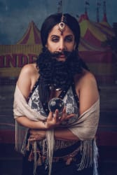Personne portant une barbe noire bouclée artificielle, un bijou sur le front, tenant une boule de cristal, avec une écharpe beige autour des épaules, devant un décor de chapiteau de cirque jaune et rouge.