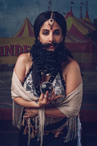 Personne portant une barbe noire bouclée artificielle, un bijou sur le front, tenant une boule de cristal, avec une écharpe beige autour des épaules, devant un décor de chapiteau de cirque jaune et rouge.