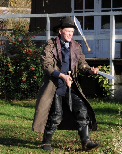 Un homme portant un chapeau noir, un long manteau marron et des bottes noires jongle avec trois machettes dans un jardin ensoleillé.