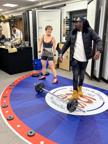 Une femme en tenue de sport rouge et noire et un homme en veste noire et bottes jaunes se tiennent sur un tapis circulaire bleu et rouge avec une barre chargée de poids au centre dans une pièce intérieure.