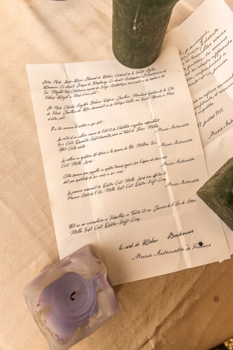 Feuille de papier avec texte manuscrit en français, posée sur une surface beige, entourée de deux bougies vertes et d'un petit porte-bougie violet.