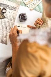 Personne écrivant une lettre avec une plume et de l'encre sur un bureau blanc, portant un vêtement ancien couleur marron clair avec dentelle blanche, vue de dessus sur l'épaule.